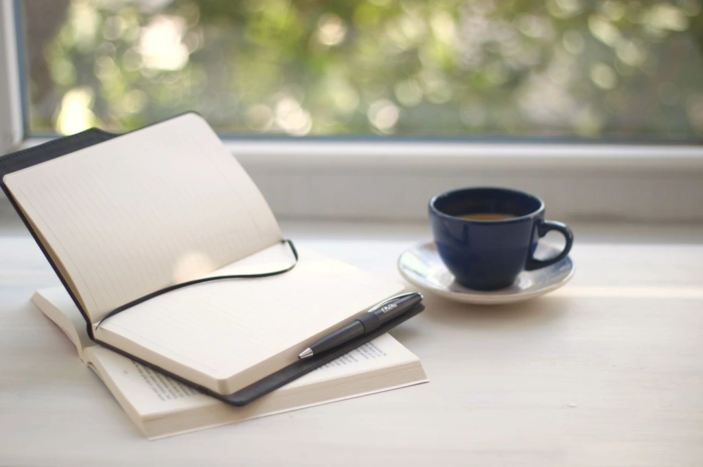 Cuaderno abierto con una pluma junto a una ventana con luz natural, para redactar el texto de una esquela